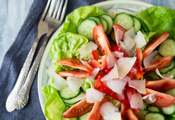 Fresh healthy salad with lettuce, olive oil and slices of tomato, cucumber, pepper and parmesan cheese on blue wooden table. Selective focus