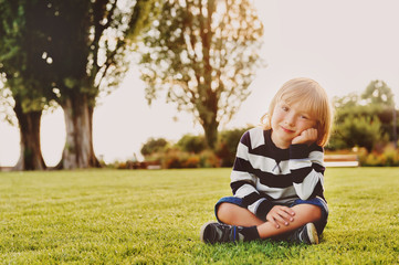 Outdoor portrait of adorable toddler boy sitting on grass at sunset