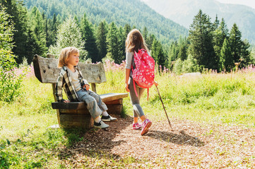 Cute little boy and his sister with backpack hiking in mountains, resting on a bench