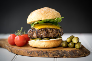 large thick grilled beef minced burger patty with toasted bun melted cheddar cheese green fresh salad cherry tomato and pickle cucumber on rustic wooden board and white table