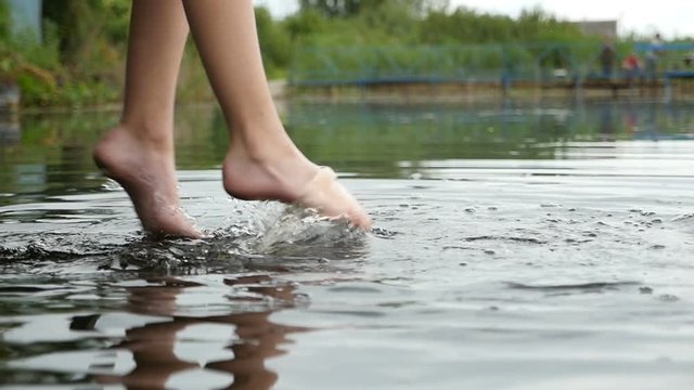 Impressive closeup  of frolicking female legs with a fashionable makeup, which are swinging, while touching water in a lake in a relaxed way. The young woman is sitting on a wooden bridge in slo-mo