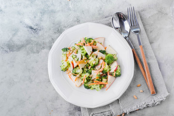 Vegetable salad with broccoli,apple,carrots, pine nuts and sauce in white plate on light stone table background. Healthy vegetarian food/ Copy space