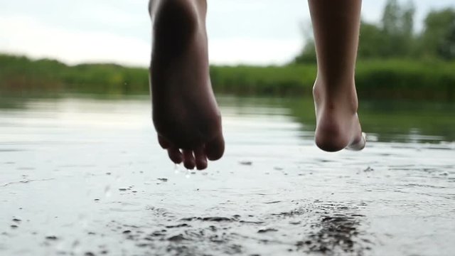 Funny closeup of slim female legs, which are swinging, while touching water in a lake in a relaxed way. The young woman is sitting on a wooden bridge in slow motion