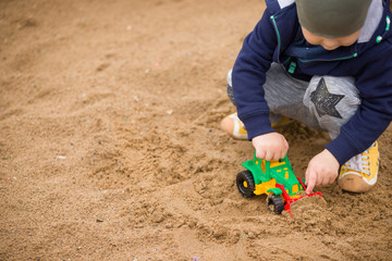 Portrait of cute toddler boy sitting on the ground and playing with toy tractor and sand in the park. Child walking outdoors. Lifestyle.