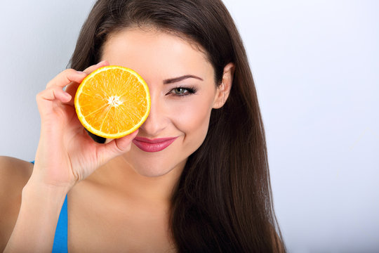 Beautiful makeup brunette woman holding fresh orange fruit near the eye and looking happy with empty space blue background. Closeup portrait