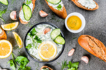 Traditional Caucasian and Greek food. Sauce tzatziki with ingredients - cucumber, lemon, parsley, dill, garlic. On a dark stone table. With sandwiches and baguette. Top view