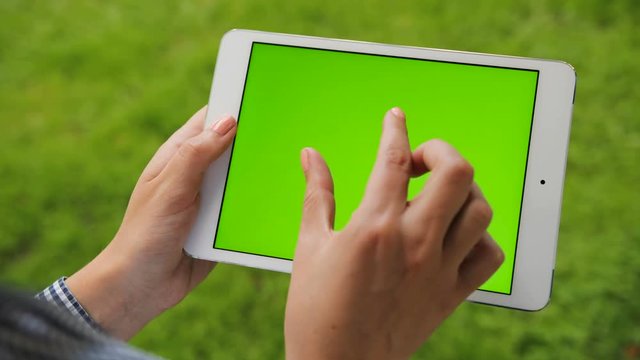Female hands holding white tablet computer device with green screen on green grass background. Outdoors in park. Woman scrolling, zooming, tapping on touchscreen. Chroma key. Close up.