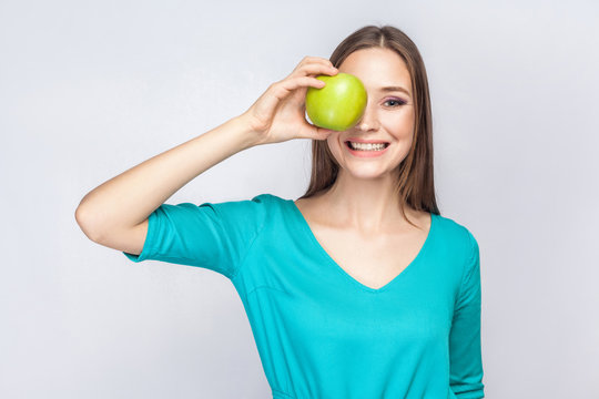 Young beautiful woman with freckles and green dress holding apple in front of her eyes and smiling . studio shot, isolated on light gray background.