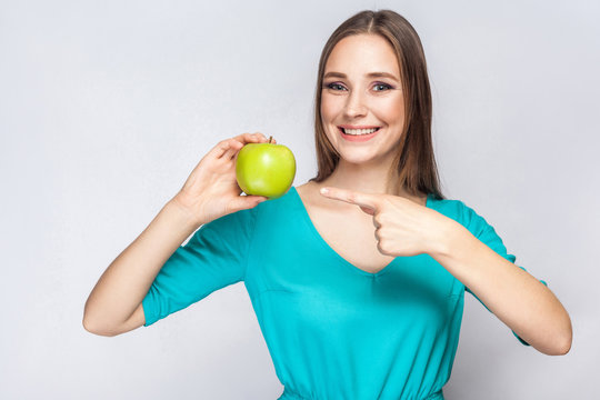 Young beautiful woman with freckles and green dress holding apple and pointing with finger. studio shot, isolated on light gray background.