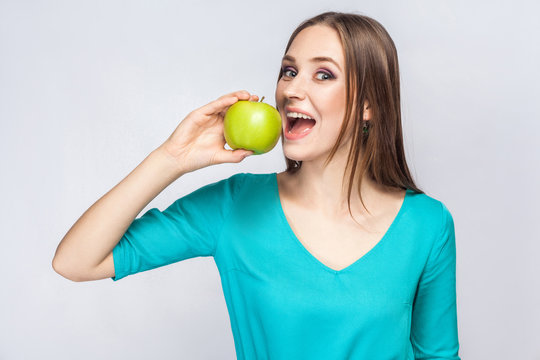 Young beautiful woman with freckles and green dress holding apple and eating looking at camera. studio shot, isolated on light gray background.