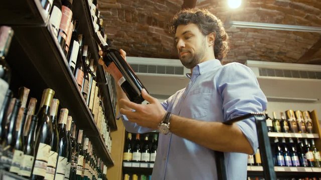 Young caucasian man with curly hair taking the bottle of wine in the supermarket and putting to the basket. Close up. View from the bottom.