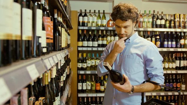Young attractive man choosing the good bottle of wine in the supermarket and putting it to the basket.