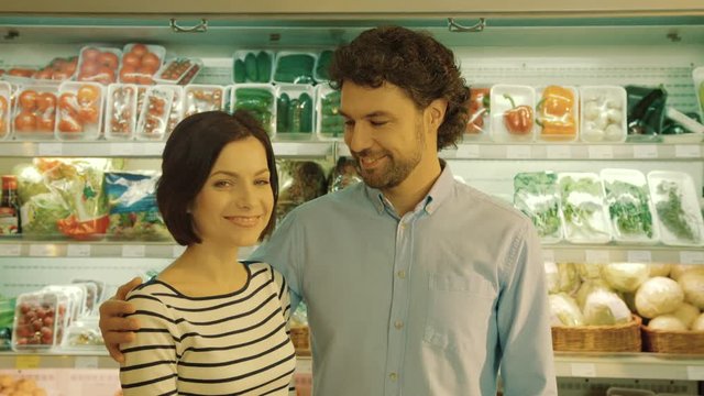Portrait of couple standing in front of the camera and smiling while do shopping in the vegetable department in the supermarket and looking to each others, feeling very happy.