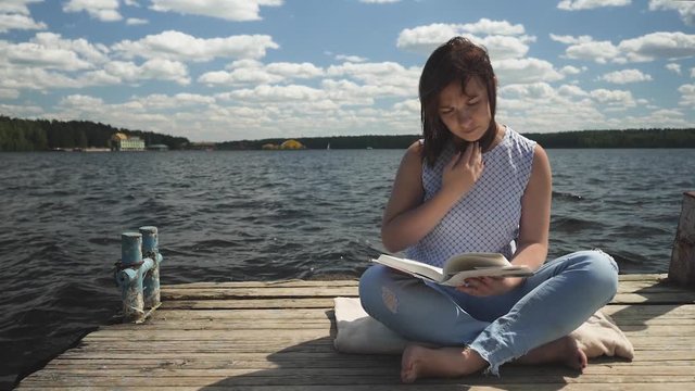 Young brunette sitting on old wooden pier in the lotus pose and reading book on background of lake and forest
