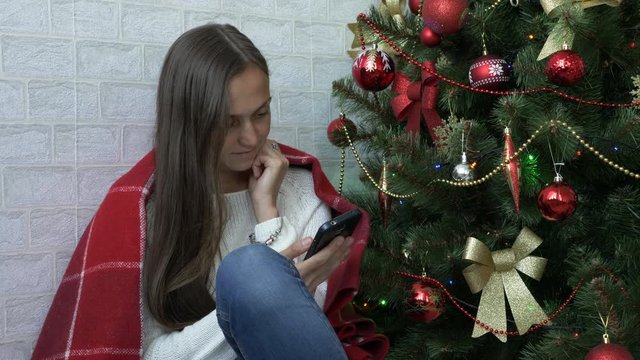 Woman sits on a floor with red plaid and using smartphone next to Christmas tree