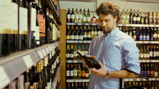 Young attractive man with curly hair taking two bottle of wine and choose on of them in the supermarket and putting to the basket. Close up.