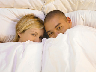 Man and woman lying in bed, holding blanket over their faces