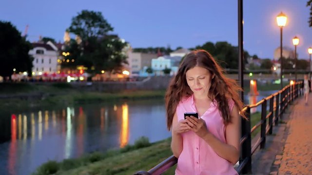 Pretty young girl using smartphone near the railing on waterfront in evening time, old city, river and bridge in the background