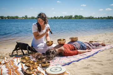 Beautiful female receiving energy sound massage with singing bowls on her body on a river bank at spring sunny day. And little black doggy interested in