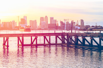 Sunset casting a pink light on a disused jetty adjacent to the Thames Barrier in London with an approaching ship and Canary Wharf in the background