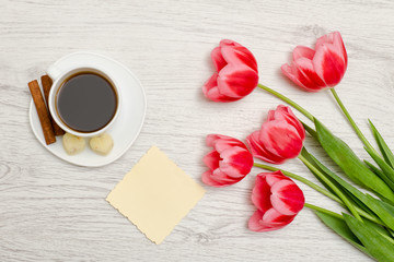 Pink tulips, blank sheet of paper, mug of coffee and cinamon, light wooden background. top view