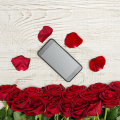 Smartphone lies on a white wooden table surrounded by petals and a bouquet of roses