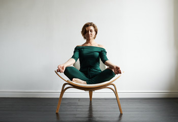 Attractive smiling young Caucasian woman in stylish jumpsuit sitting on chair in lotus pose in...