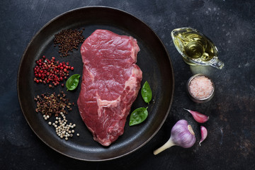 Above view of a frying pan with raw fresh ribeye beefsteak and various seasonings on a dark metal background, horizontal shot