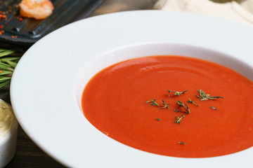Red tomato soup gazpacho in a large white plate on a table decorated with ingredients. Macro closeup view.