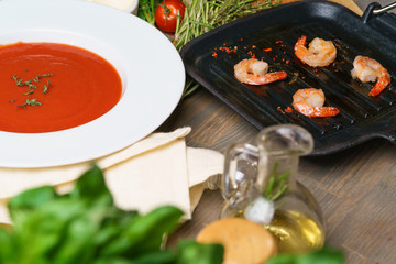 Red tomato soup gazpacho in a large white plate on a table decorated with ingredients. Leaves of basil and rosemary, fried prawns on a black frying pan, cherry tomatoes, croutons and parmesan cheese.