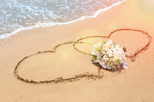 Bridal bouquet and hearts drawn on sand near water. Wedding concept