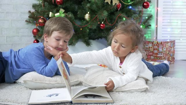 Two little cute siblings reading a book on the floor near Christmas tree