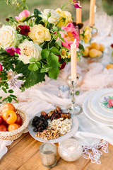 picnic, food, summer, holiday concept - close up on the festively decorated table with beautiful bouquet of roses and pink peonies, candles in candlesticks, dried fruits, nectarines, nuts, glassware