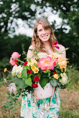 wedding, celebration, beauty, country life, spring, nature concept - young attractive woman with light brown hair in summer dress with floral print puting out nice bunch of various flowers