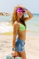 Beautiful sporty woman in bikini, jeans shorts and transparent cap look to the camera. Summer holidays, vacation on a tropical island, clear water of ocean and hot sand. Portrait of a sexy young lady.