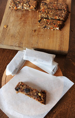 Protein muesli bar with nuts and dried fruits on baking parchment paper in the process of wrapping. Three wrapped. Large uncut plank in the background on a wooden board