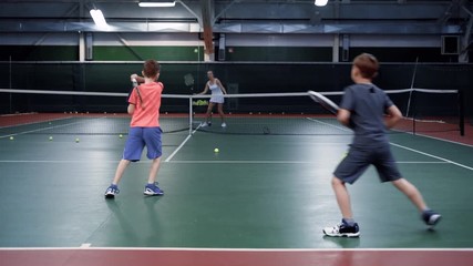 A female trainer in a sports suit teaches playing tennis in two middle-aged boys, guys are bouncing balls on a tennis court