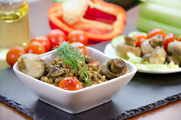 Dish from buckwheat with baked mushrooms, tomatoes, pepper. Decorated with a branch of fresh dill. In the background, a grease cup with olive oil, fresh red sweet pepper, celery. Home dietary cuisine.