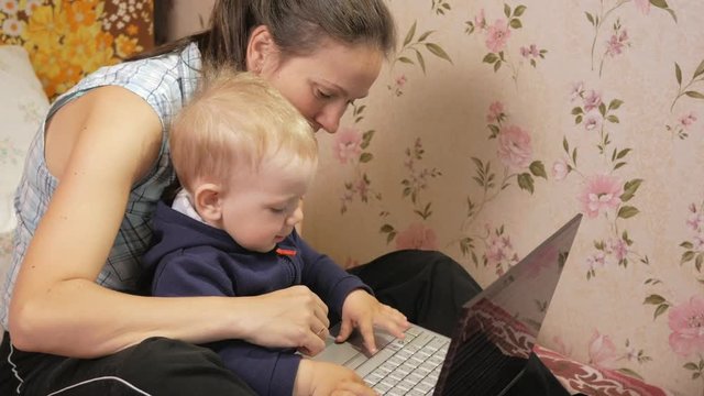 Young woman with her son sitting at home on the couch with a laptop. Look carefully at the screen and press the keys.