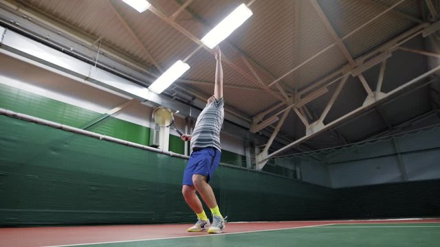 An adult male in sports clothes serves a serve with a racket and a tennis ball, the person is in a closed stadium of a sports complex