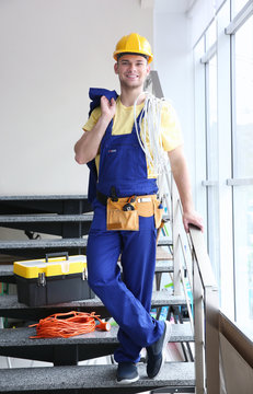 Smiling young electrician with bunch of wires and toolbox standing on stairs indoors