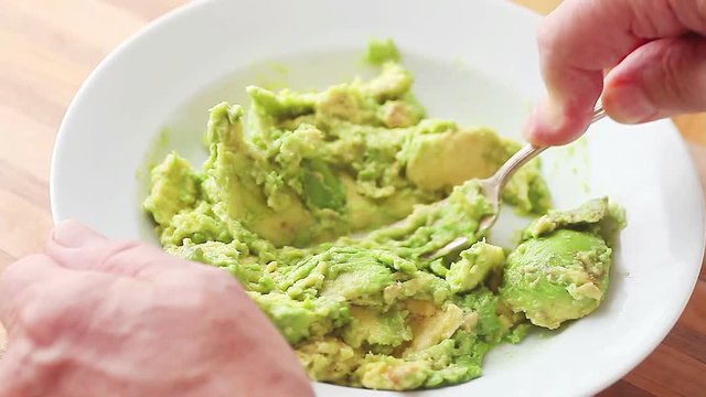 A man uses a fork to mash an avocado in a white dish