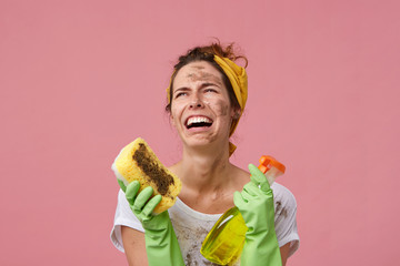 Hopeless crying housewife with dirty clothes and face wearing casual clothes and gloves holding...