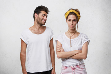 Studio shot of handsome positive bearded male in white t-shirt trying to convince or apologize to his angry upset girlfriend in yellow headband who is looking offended, keeping her arms crossed - obrazy, fototapety, plakaty