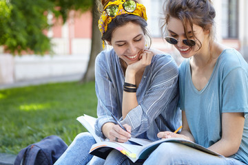 Happy smiling females sitting outdoors reading books preparing for classes at university having fun...
