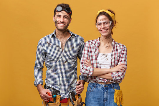 Indoor portrait of positive cute young couple of professional maintenance workers wearing overalls and protective eyeglasses smiling cheerfully, enjoying helping people fixing household problems