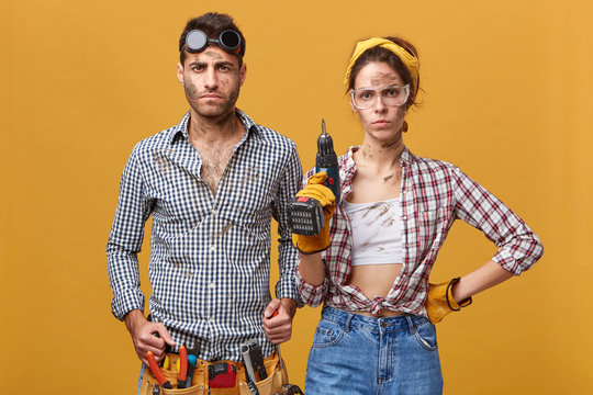 Indoor shot of confident young maintenance workers or repairmen in overalls, protective wear, standing at studio wall equipped with tools and instruments, ready for doing repairs and fixing things