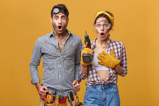 Beautiful female electrical technician with drill wearing protective gloves and goggles standing next to her attractive male colleague with belt kit of tools, both staring at camera with shocked looks