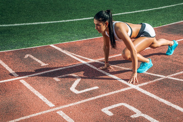 A young athletic woman stands in a low start for running in the stadium on a summer day