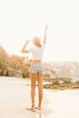 Beautiful fit lady in white shirt and shorts goes along the line of waves and touch cap in sunset rays of light. Beauty sunshine cute girl on a tropical sand beach sea ocean shore with large stones.